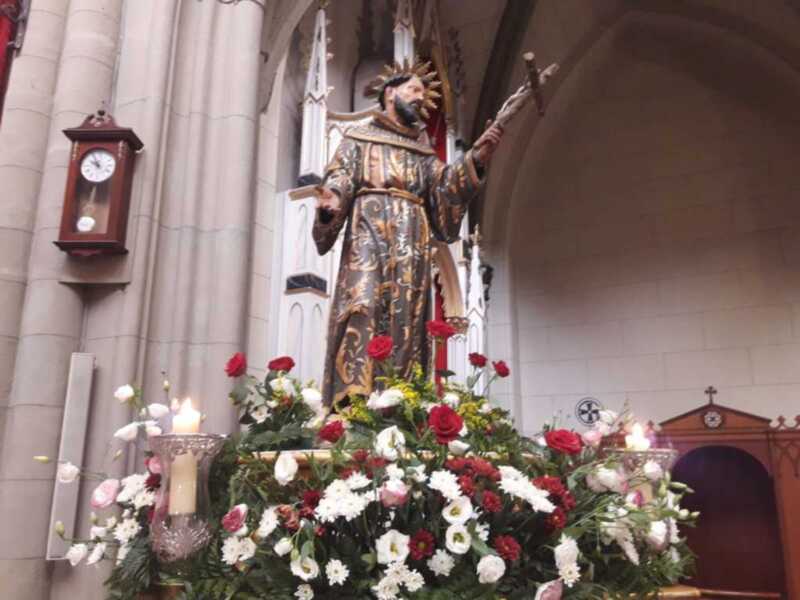 Una estatua de un rey vestido con armadura dorada y corona, rodeada por flores y velas encendidas en una iglesia gótica.