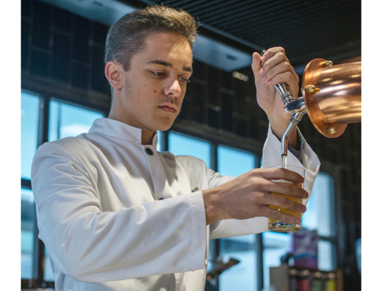 Un barman en uniforme blanco prepara una bebida con un tapon de cobre en un entorno moderno con grandes ventanales.