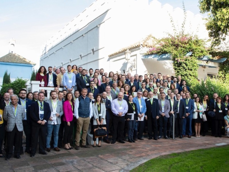 Grupo de personas posando para una foto en un evento formal, con edificios y vegetación en el fondo.