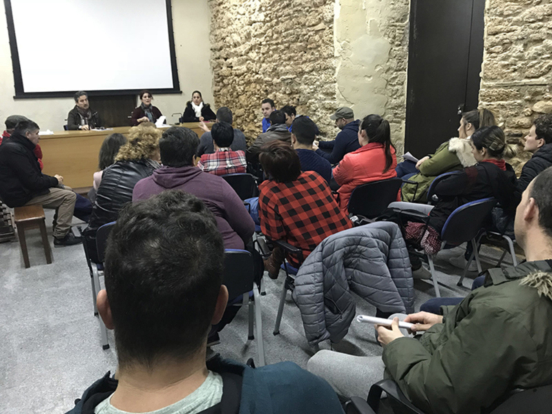 Una audiencia en una sala con paredes de piedra, donde un grupo de personas se sienta en sillas, escuchando a los ponentes en una mesa de conferencia.