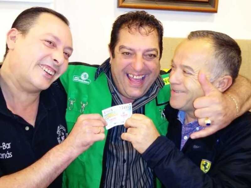 Tres amigos sonrientes celebrando con un billete de lotería en una sala acogedora.