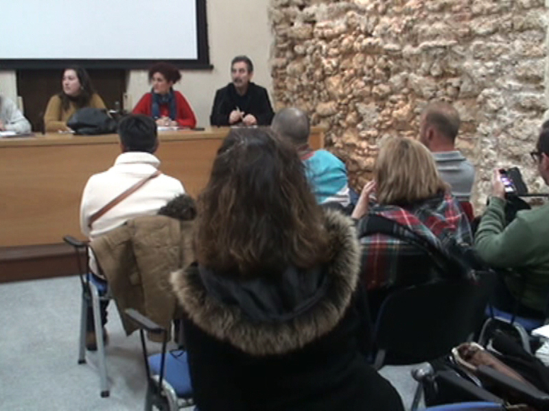 En una sala de conferencias con paredes de piedra, un grupo de personas se encuentra sentado en sillas y una persona está tomando notas. Al fondo, hay un panel de presentación con tres personas sentadas en una mesa. La escena sugiere que se está celebrando un evento académico o de conferencia.