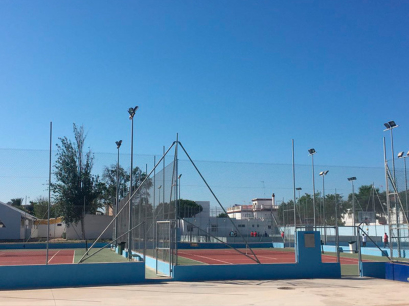Cancha de tenis con redes y postes altos, bajo un cielo azul claro. La cancha está rodeada por una cerca y hay árboles en el fondo.
