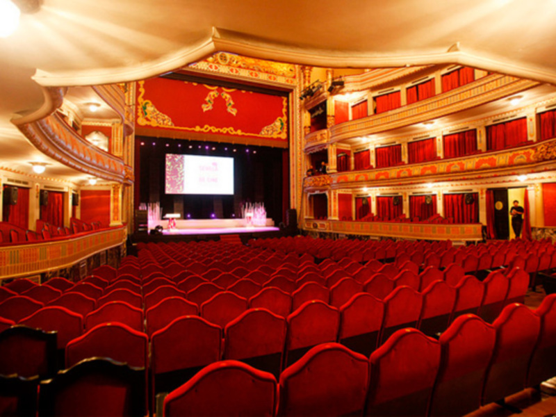 Interior de un teatro con asientos rojos y una pantalla grande en el escenario. El teatro está iluminado con luces blancas y la decoración es elegante con detalles dorados en el techo y la pantalla.
