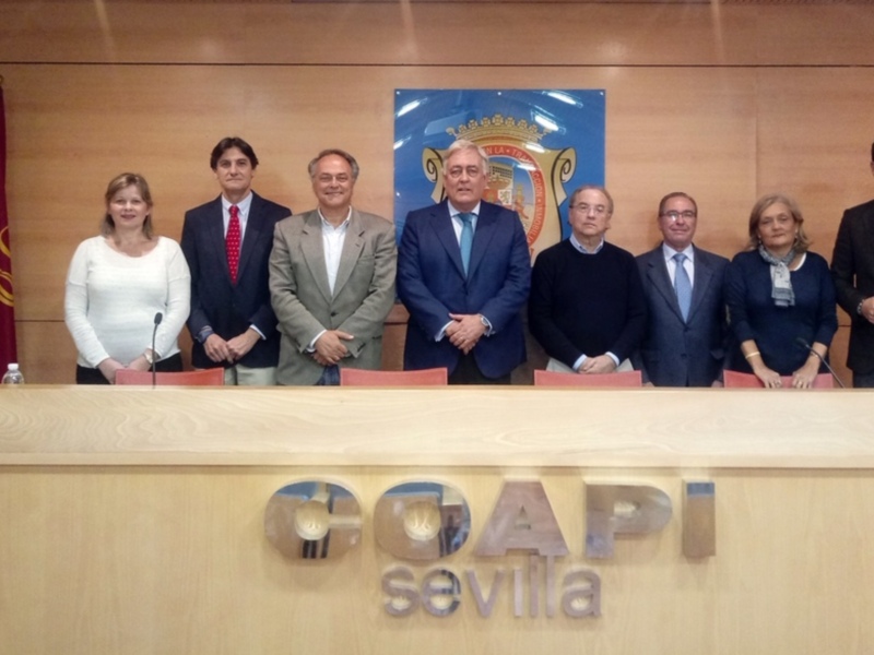 A una fila de personas posando frente a un escenario con el logo de la CAPII Sevilla. La persona en el centro lleva una chaqueta azul y es la figura más destacada. A su izquierda, hay dos personas con camisas blancas y a la derecha, cuatro más con camisas de colores diferentes. En el fondo, se pueden ver dos banderas y una pancarta con la imagen de un escudo. La escena parece ser parte de una ceremonia o evento oficial en Sevilla, España.