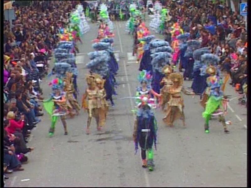 Desfile de carnaval con participantes en trajes coloridos y decorativos, observado por una multitud en la acera.
