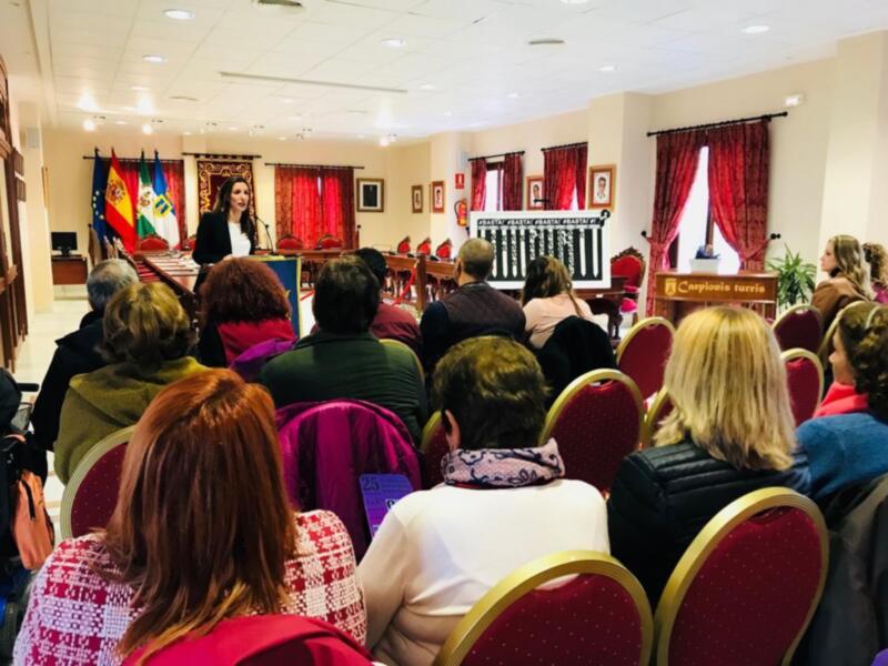 A una mujer hablando en un evento público. La sala está decorada con banderas y cortinas rojas, y hay asistentes sentados en sillas rojas. La mesa central tiene un cartel informativo.