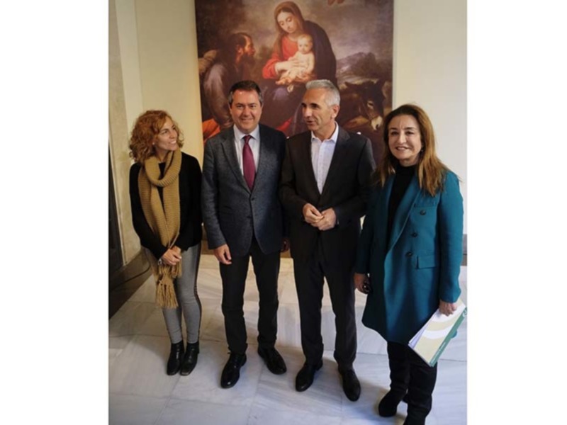 Una imagen de tres personas posando frente a un lienzo artístico con una escena religiosa. La persona en medio lleva un saco de lana y una camisa blanca, mientras que la persona a su derecha sostiene un cuaderno. La escena sugiere una reunión formal o de trabajo en un entorno cultural.