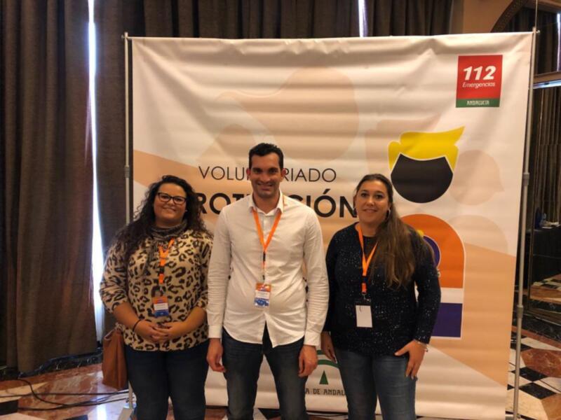 Tres personas posan frente a un banner que dice "Voluntariado 112 Emergencias". La imagen parece ser de un evento o conferencia relacionado con la emergencia y voluntariado.