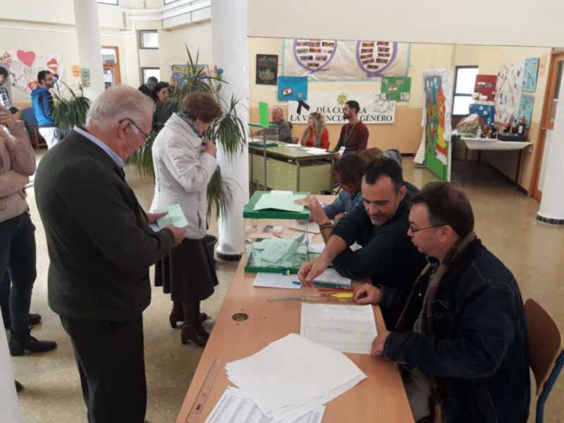 Votación en un colegio electoral con personas votando y funcionarios de mesa.