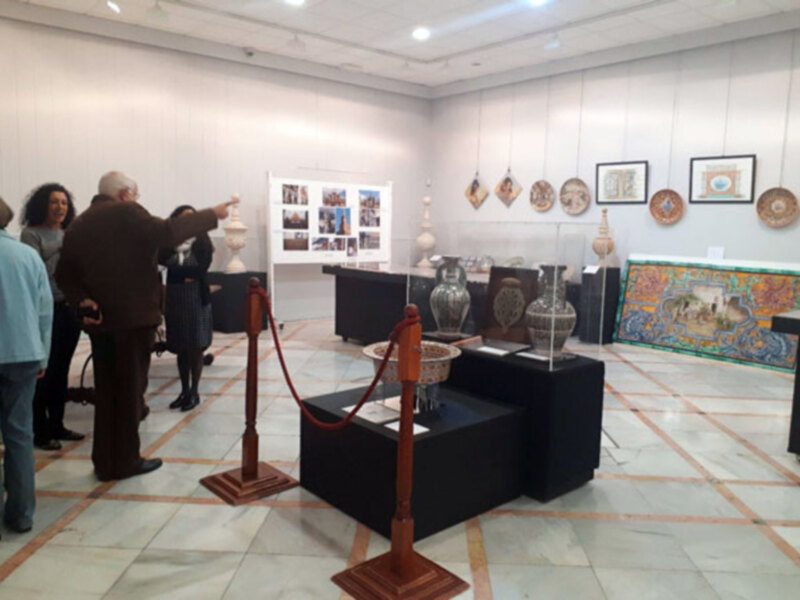Una sala de exposiciones con vitrinas y objetos artísticos colgados en la pared. Personas observando el contenido de las vitrinas y conversando entre sí.