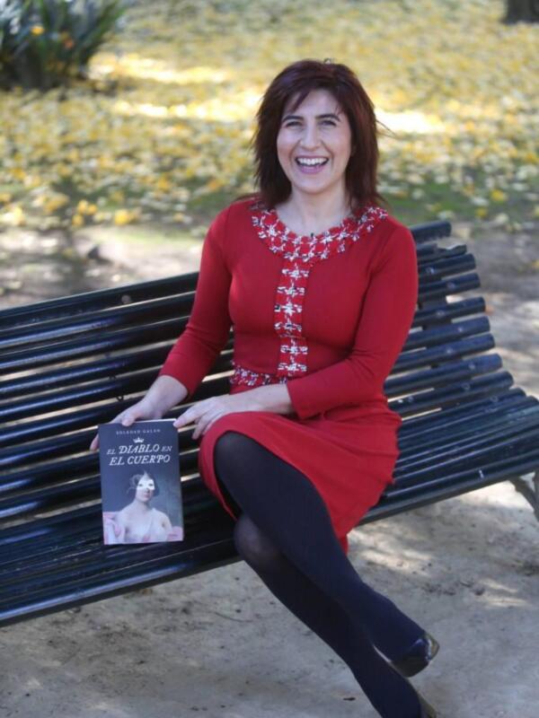 Una mujer sonríe ampliamente sentada en un banco de parque, rodeada por hojas amarillas caídas. Lleva un vestido rojo con detalles bordados y pantalones negros. A su lado, hay un libro titulado "El Diario de El Cuerpo".