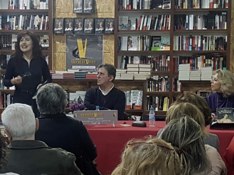 Una mujer habla en un escenario frente a una audiencia, con libros y fotografías en el fondo. La imagen muestra un ambiente de lectura y debate.