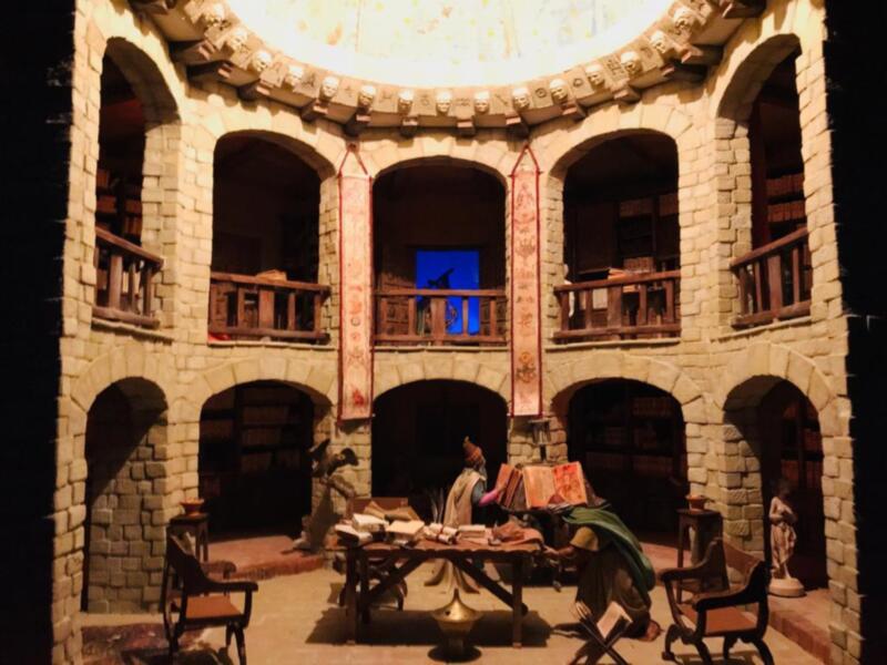 Un interior medieval de piedra con arcos y balcones, donde dos personajes se sientan en una mesa baja. La atmósfera es histórica y dramática, con detalles como un techo dorado y una figura en la parte superior.