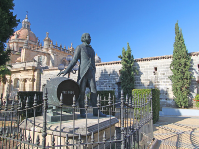 Escultura de un hombre en el centro, rodeada por un muro de piedra y un árbol. En el fondo se puede ver una catedral con cúpulas y torres. El cielo es azul claro, indicando un día soleado.