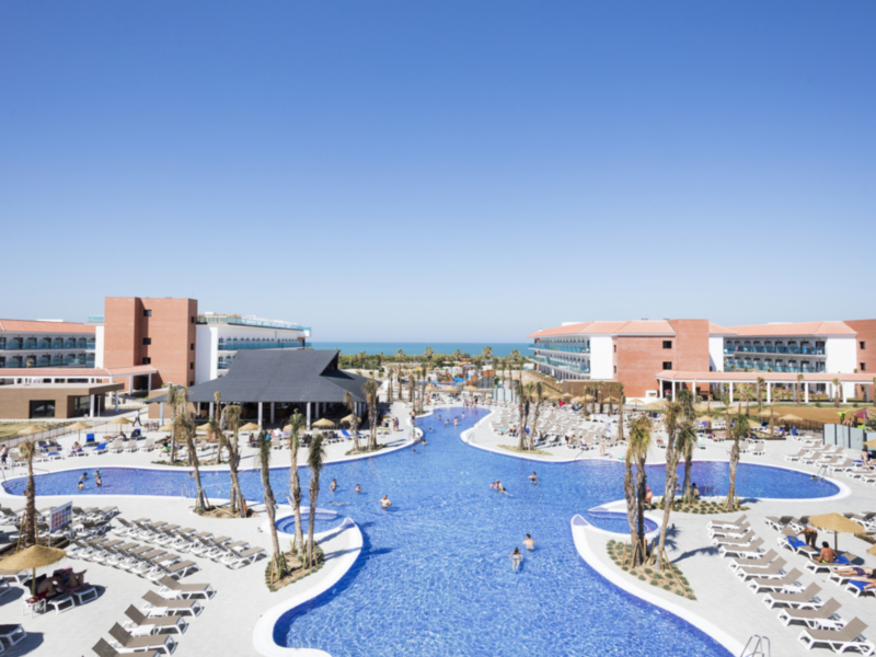 Hotel con piscina y playa frente al mar en un día soleado.