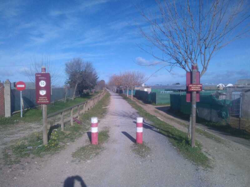 Un camino de tierra sinuosamente curvado, flanqueado por una cerca de madera y un poste con un cartel rojo. Al fondo, se observa un cielo azul claro con algunas nubes dispersas y árboles sin hojas.