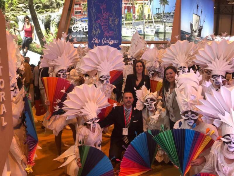 Una multitud de personas con disfraces de calaveras y sombreros de plumas, posando frente a un fondo con una pantalla gigante que muestra imágenes de barcos y paisajes.