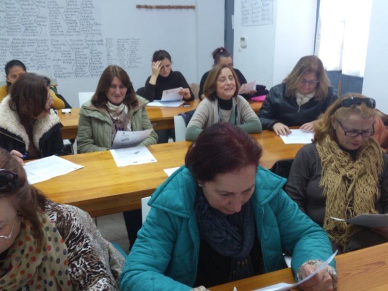 En una aula con paredes blancas, varias personas están sentadas en mesas de madera, escribiendo en cuadernos. La imagen muestra un ambiente de estudio o clase.