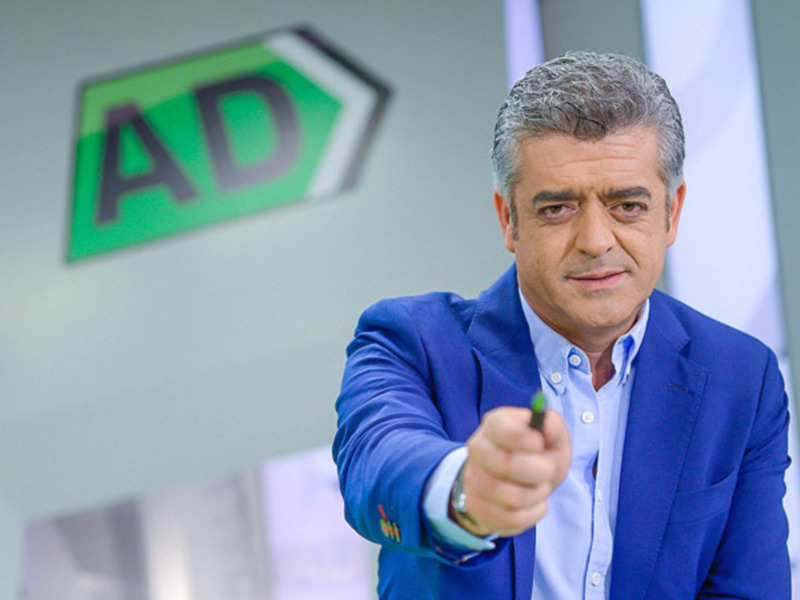 Un hombre en un traje azul apuntando con una pluma verde. En el fondo, se puede ver un logotipo de AD.