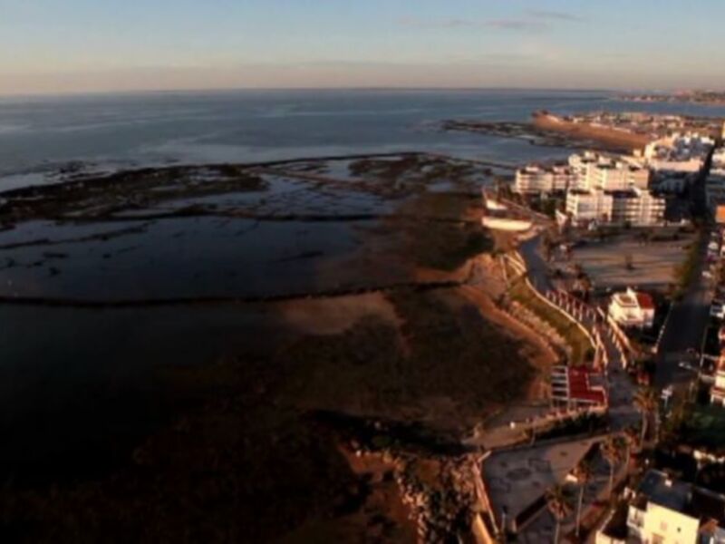 Imagen aérea de una ciudad costera con edificios blancos y verdes, rodeada por el mar en la puesta del sol.