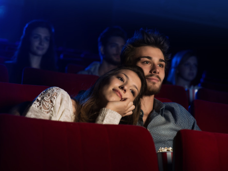 Pareja disfrutando de una película en el cine, sentados en un asiento rojo bajo la iluminación dramática.