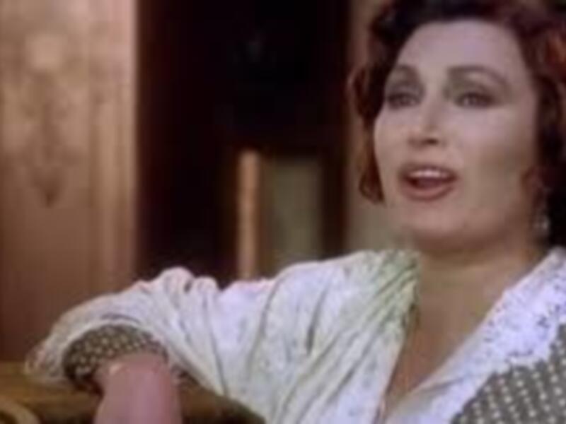 Una mujer con cabello rizado y vestido con detalles de encaje, posando con expresión de sorpresa o sorpresa.