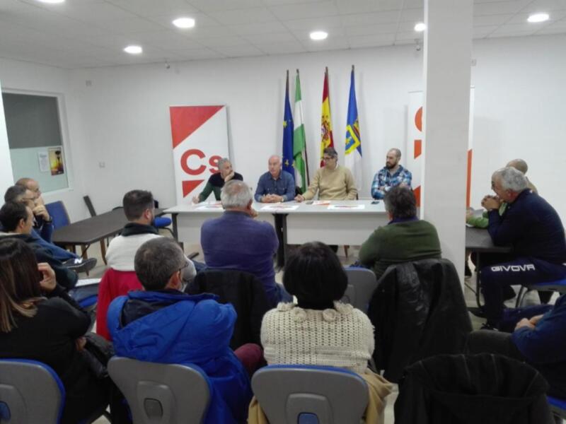 Una reunión de trabajo en un salón con varias personas sentadas alrededor de una mesa, banderas y carteles en el fondo.