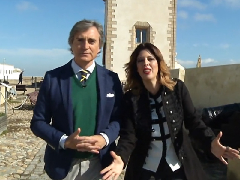 Un hombre y una mujer en un entorno urbano, posiblemente en una ciudad histórica. El hombre lleva un traje azul con camisa blanca y corbata, mientras que la mujer usa una chaqueta negra con detalles blancos. Ambos parecen estar en un entorno arqueológico o histórico, posiblemente en una ciudad antigua.