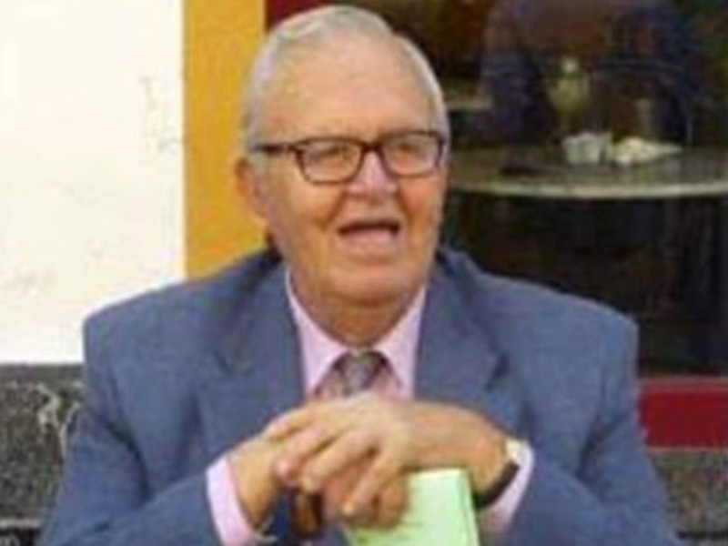 Un hombre mayor con cabello blanco, gafas y un traje azul, sosteniendo un libro en una mesa con una taza de café y una servilleta.