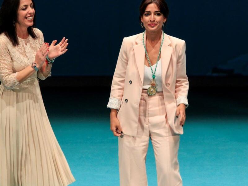 Dos mujeres en un evento, una con un vestido blanco y la otra con un traje beige, sonríen y aplauden. Fondo azul con iluminación de escenario.