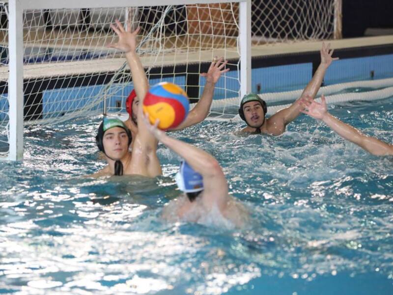 Un grupo de jugadores de waterpolo en acción, con un balón rojo y amarillo visible entre ellos. El agua es cristalina y el objetivo parece ser un gol en una portería con redes blancas.