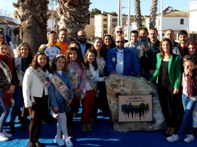 Una agrupación de personas posando frente a una placa con el nombre "CLOMITA" y un dibujo de la ciudad. La imagen muestra una escena festiva en un entorno urbano con palmeras y edificios al fondo.
