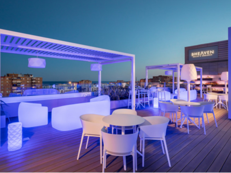 Terraza de un club nocturno con mesas y sillas iluminadas con luces de color azul, en una noche estrellada con el mar al fondo.