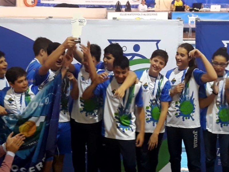 Un grupo de jóvenes jugadores de fútbol celebran su victoria, sosteniendo una bandera y medallas. El ambiente es festivo y lleno de orgullo, reflejando el éxito del equipo.