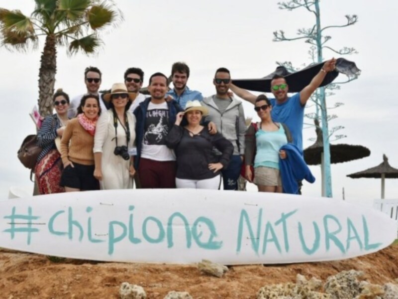 Fotografía de un grupo de personas posando con una tabla de surf en la playa, con el hashtag #chilpiono Natural.