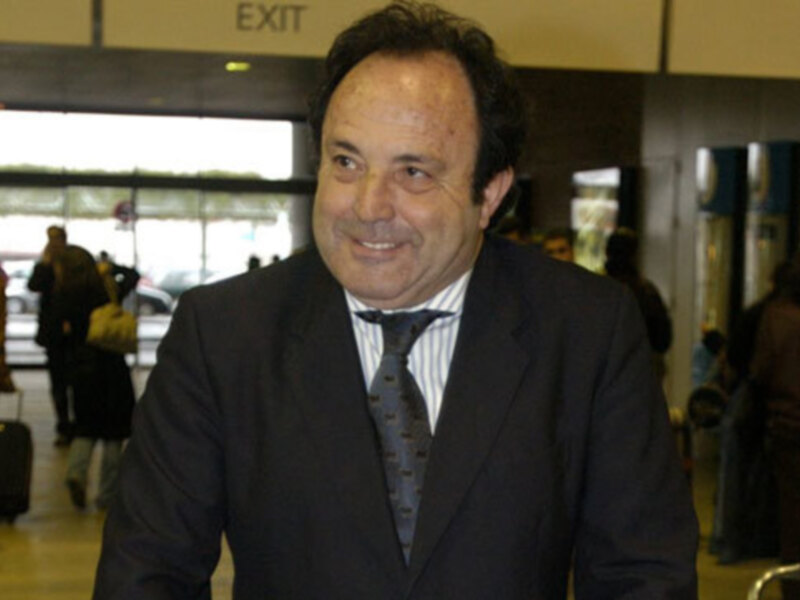 Hombre sonriente en traje y corbata en un área de viaje con personas y equipaje al fondo.