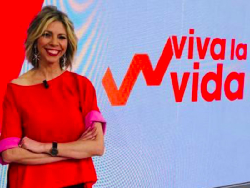 A una mujer con el cabello corto y rubio, sonriendo ampliamente. Lleva un vestido rojo con mangas de color rosa y una correa en el hombro. En el fondo, hay un logo con la palabra "Viva" y el nombre de una cadena televisiva.