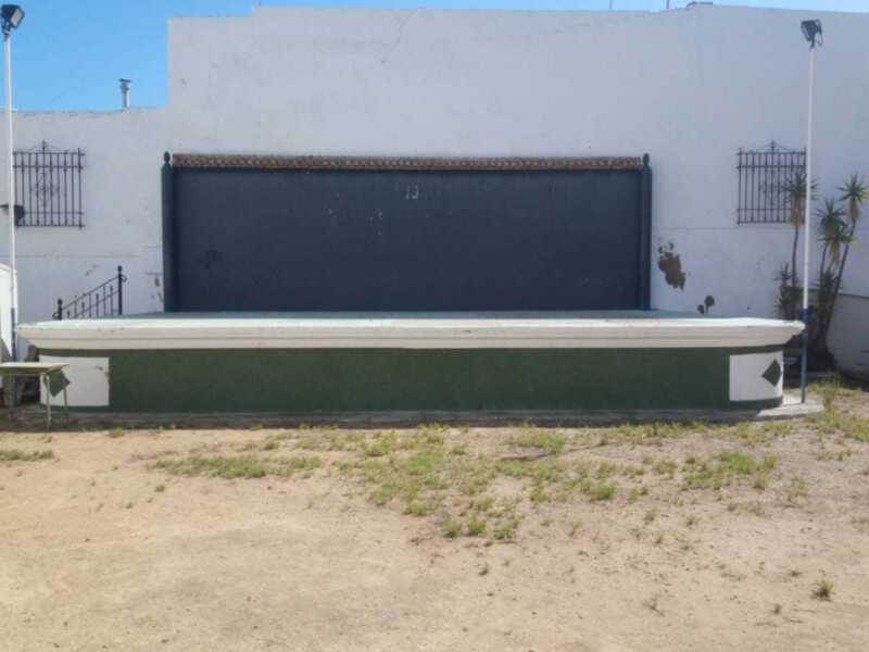 Un gran garaje de color azul oscuro con una puerta negra está ubicado en la parte superior de un edificio blanco. La imagen también muestra una estructura metálica blanca y verde con detalles negros, que parece ser parte de un sistema de riego o una estación de bombeo. La escena se encuentra en un entorno abierto con tierra seca y algunos arbustos verdes.
