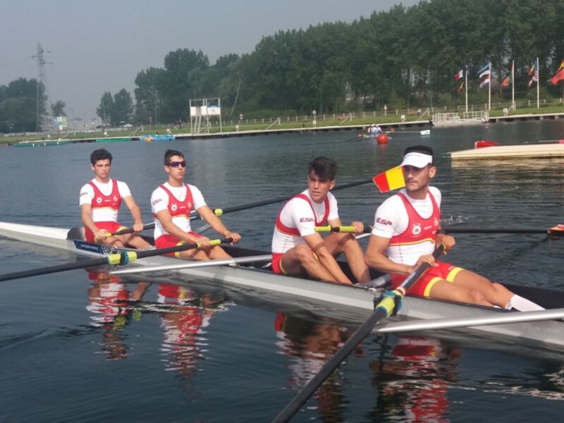 Cuatro personas remando en una canoa en un lago con banderas en el fondo.