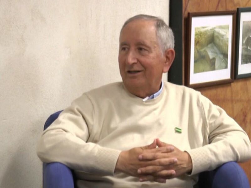 Un hombre mayor con una camiseta blanca y un cuello azul está sentado en una silla de color azul. Sus manos están juntas sobre su pecho, y detrás de él hay dos cuadros enmarcados que parecen mostrar imágenes arqueológicas. La pared de fondo es blanca y la madera en el lado derecho del cuadro da una sensación de espacio.
