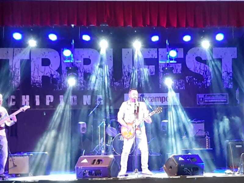 Tres músicos en un escenario con luces de neón. La palabra "BRUJIFEST" se puede leer en el fondo, junto con la frase "CHIPO".