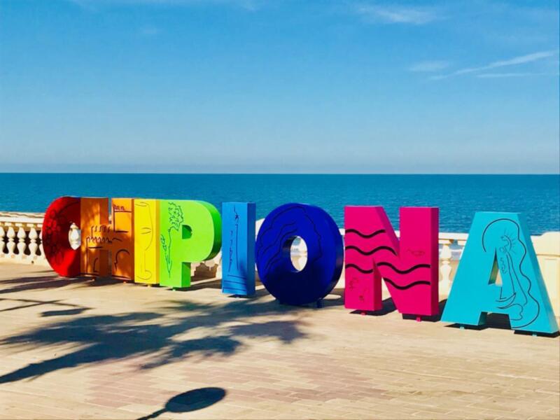 Pié de foto: Letras multicolores que forman la palabra "CIPIONA" en una playa con el mar y cielo azul al fondo.