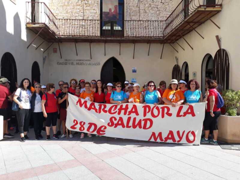 Mujeres y hombres con camisetas coloridas sostienen un cartel blanco con letras rojas que dice "MARCHA POR LA SALUD 28 DE MAYO". La imagen se toma frente a un edificio de piedra con balcones y ventanas.
