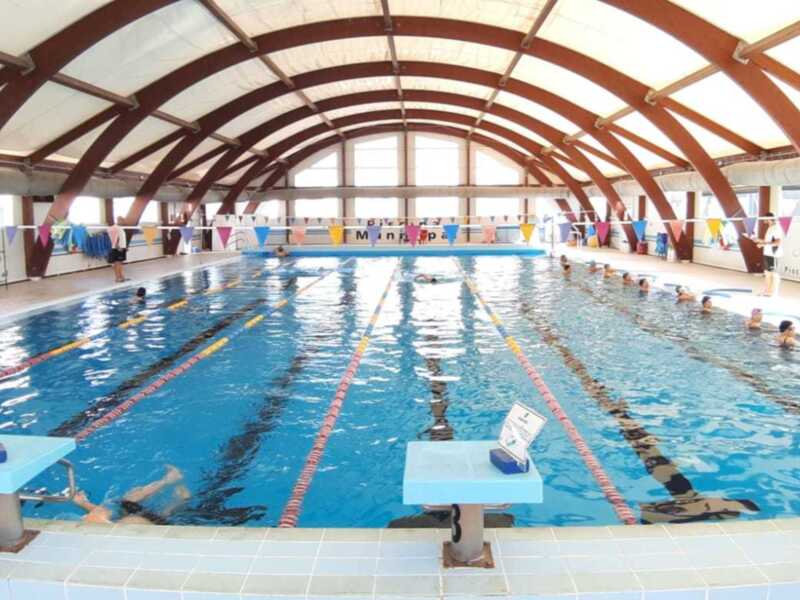 Un interior de piscina con natación, paredes de cristal y techo curvo.