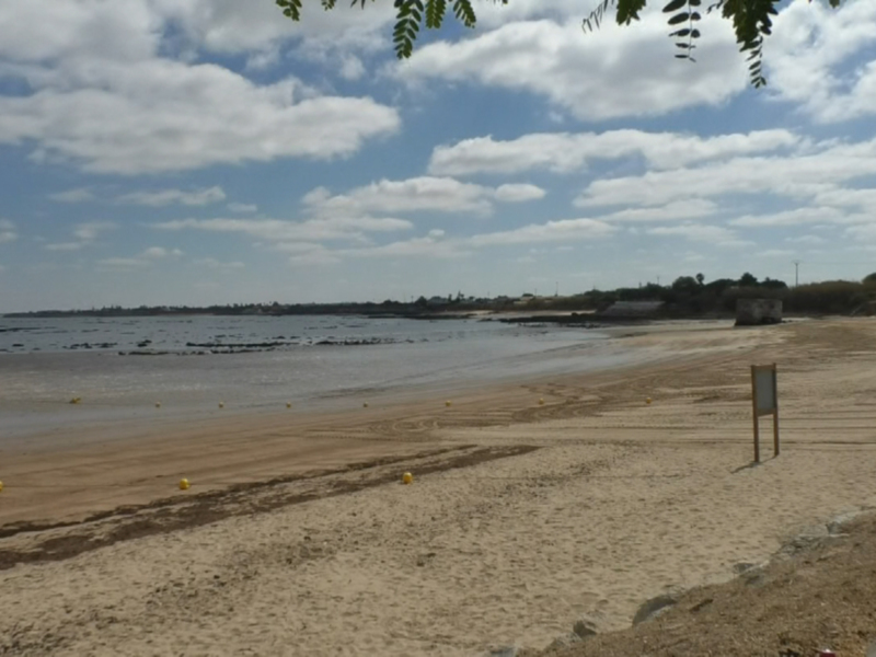 Playa tranquila con arena dorada, agua azul y nubes blancas. Señal de dirección en el extremo derecho.