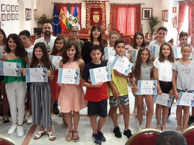 Un grupo de niños y niñas están posando para una foto en un salón. Algunos niños llevan camisetas con dibujos, mientras que otros sostienen carteles. La decoración incluye una bandera española y cortinas rojas, con plantas en el fondo. La imagen transmite alegría y orgullo escolar.
