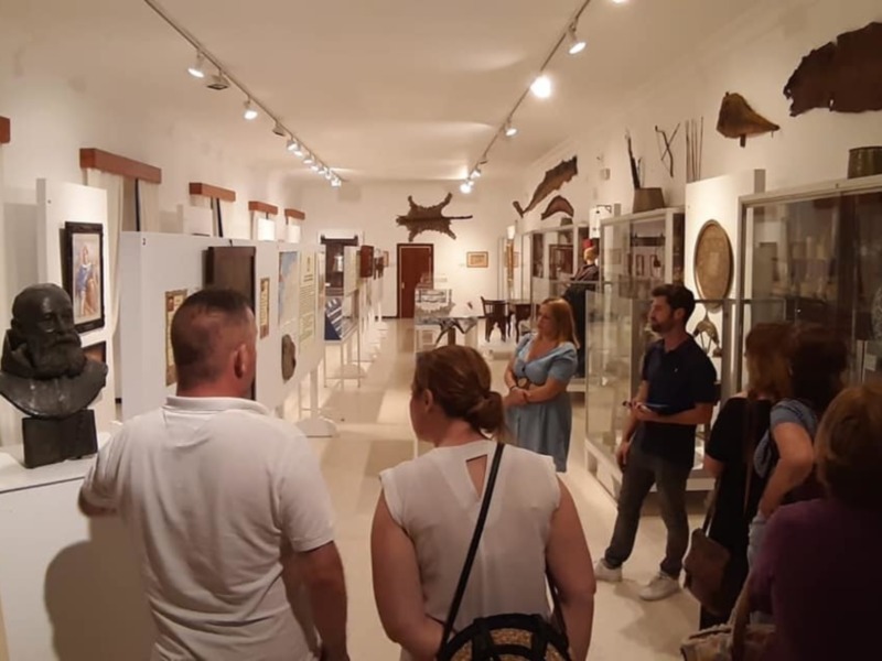 Gente visitando una galería de arte moderno. La pared está decorada con obras de arte y vitrinas con objetos antiguos. La iluminación es suave, creando un ambiente tranquilo y reflexivo.