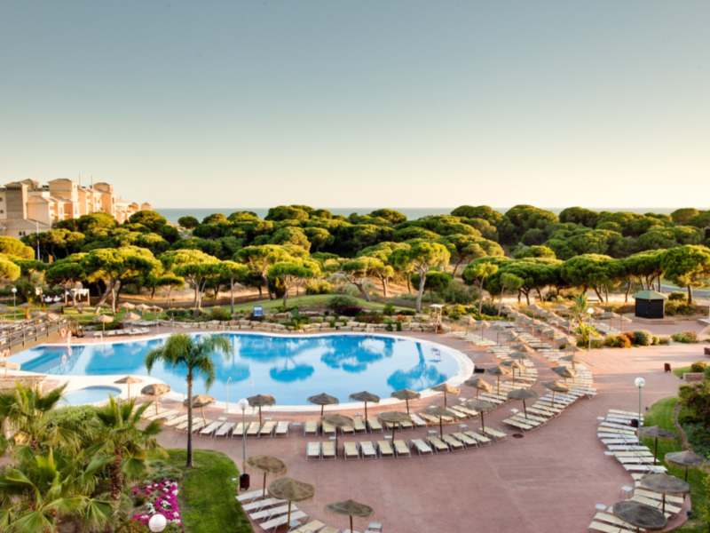 Un hermoso resort con una piscina grande y azul, rodeada de palmeras y sombrillas. La vista se extiende hasta el mar, con edificios en la distancia y un cielo claro.