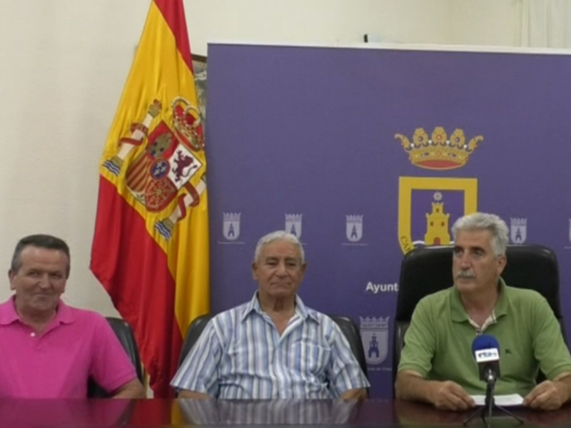 Tres personas sentadas en una mesa con una bandera española y un escudo de armas en el fondo.